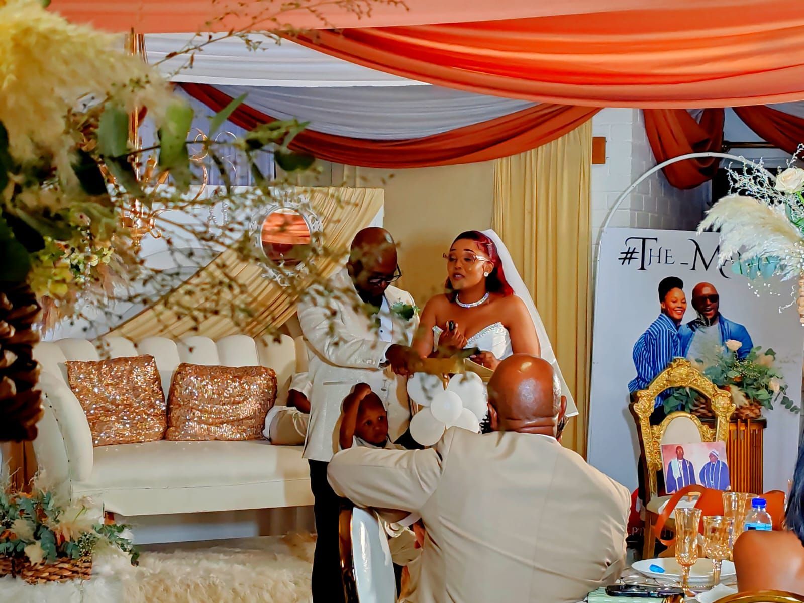 Wedding couple posing on a decorated stage with orange drapes, flowers, and a white sofa