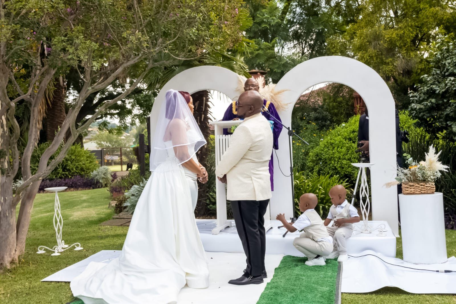 Outdoor wedding ceremony under a white arch on a lawn, with the bride and officiant standing face-to-face.