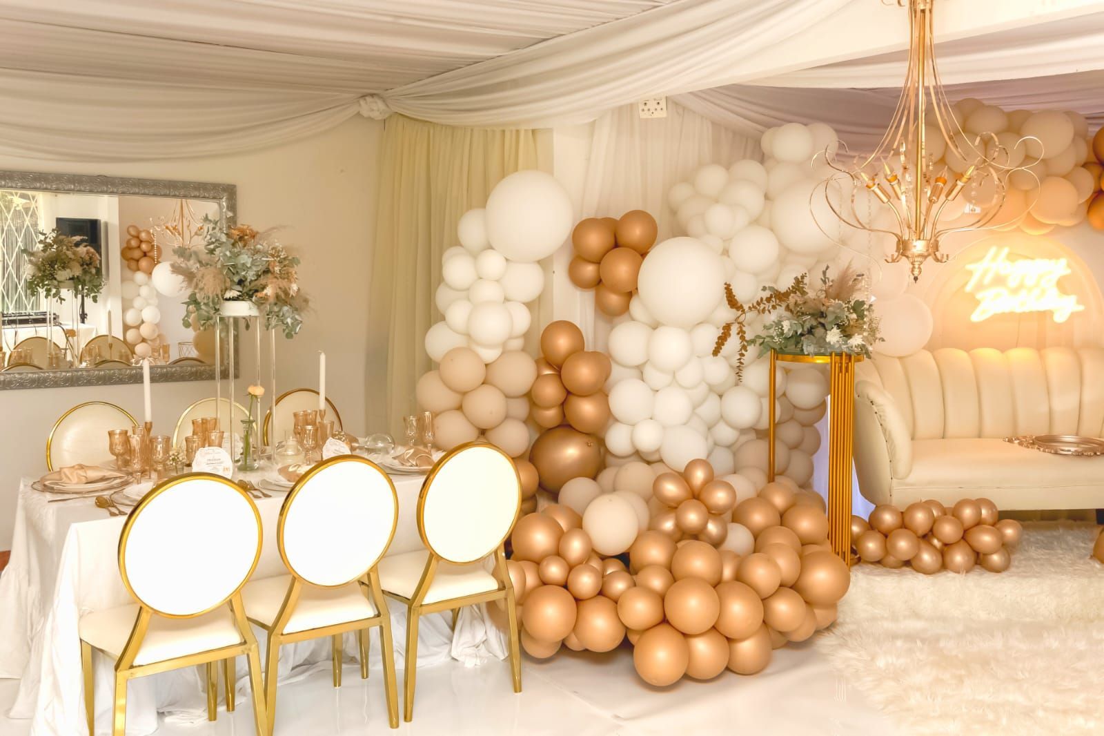 Elegant party setup with gold chairs, white and rose-gold balloons, and a cream lounge backdrop