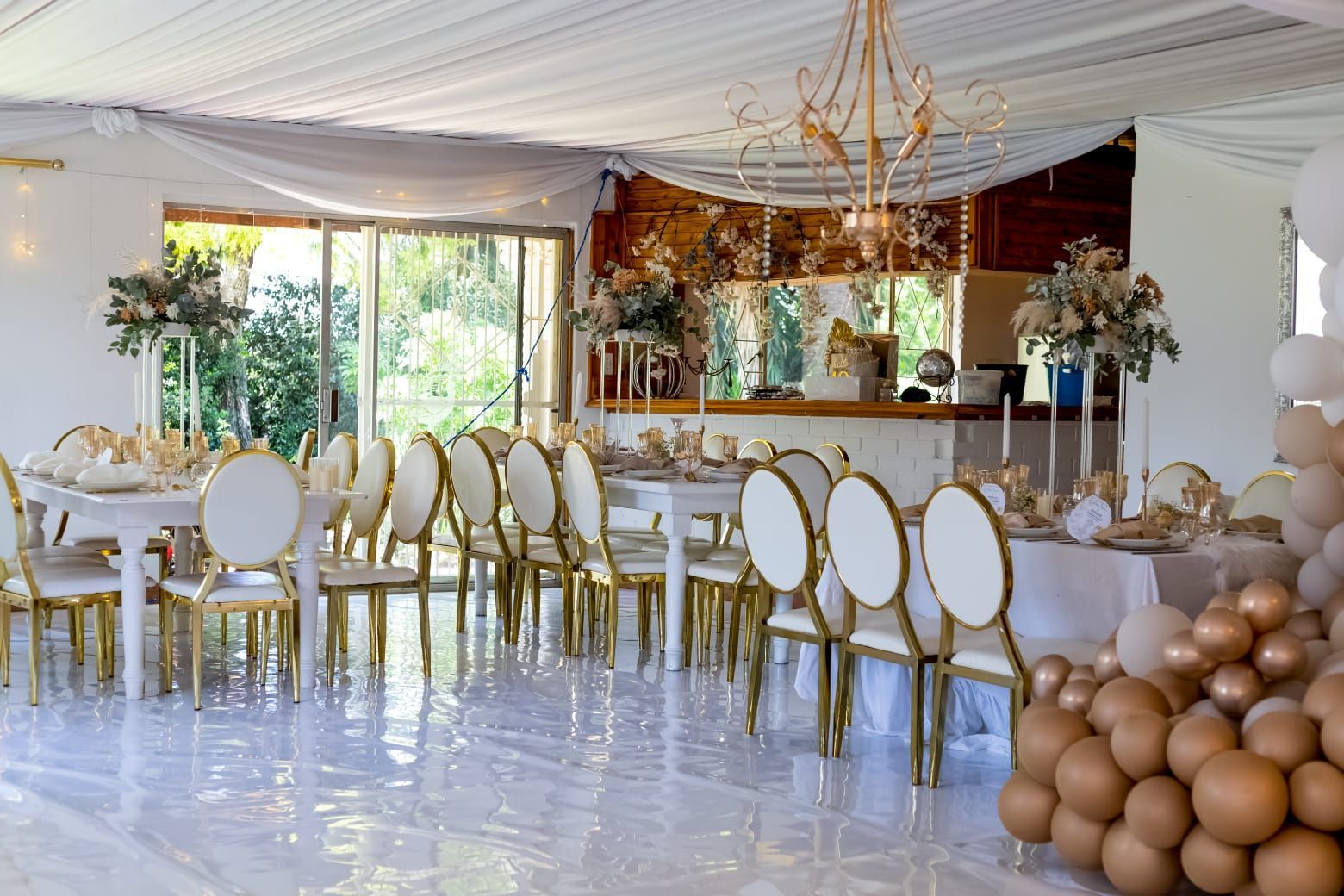 Elegant event hall with white and gold chairs, mirrored tables, and decorative balloons under a draped ceiling.