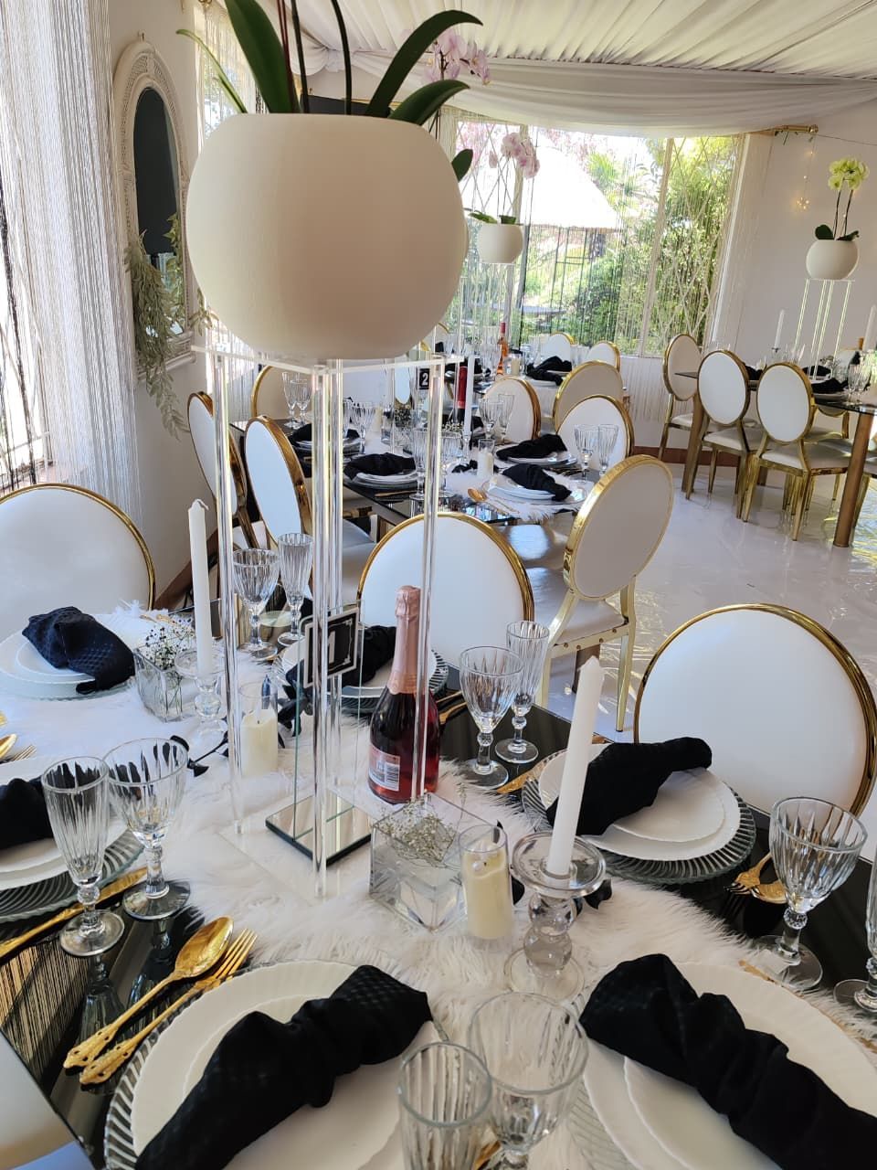 Elegant banquet table set with gold chairs, white decor, black napkins, and tall centerpiece in a bright room