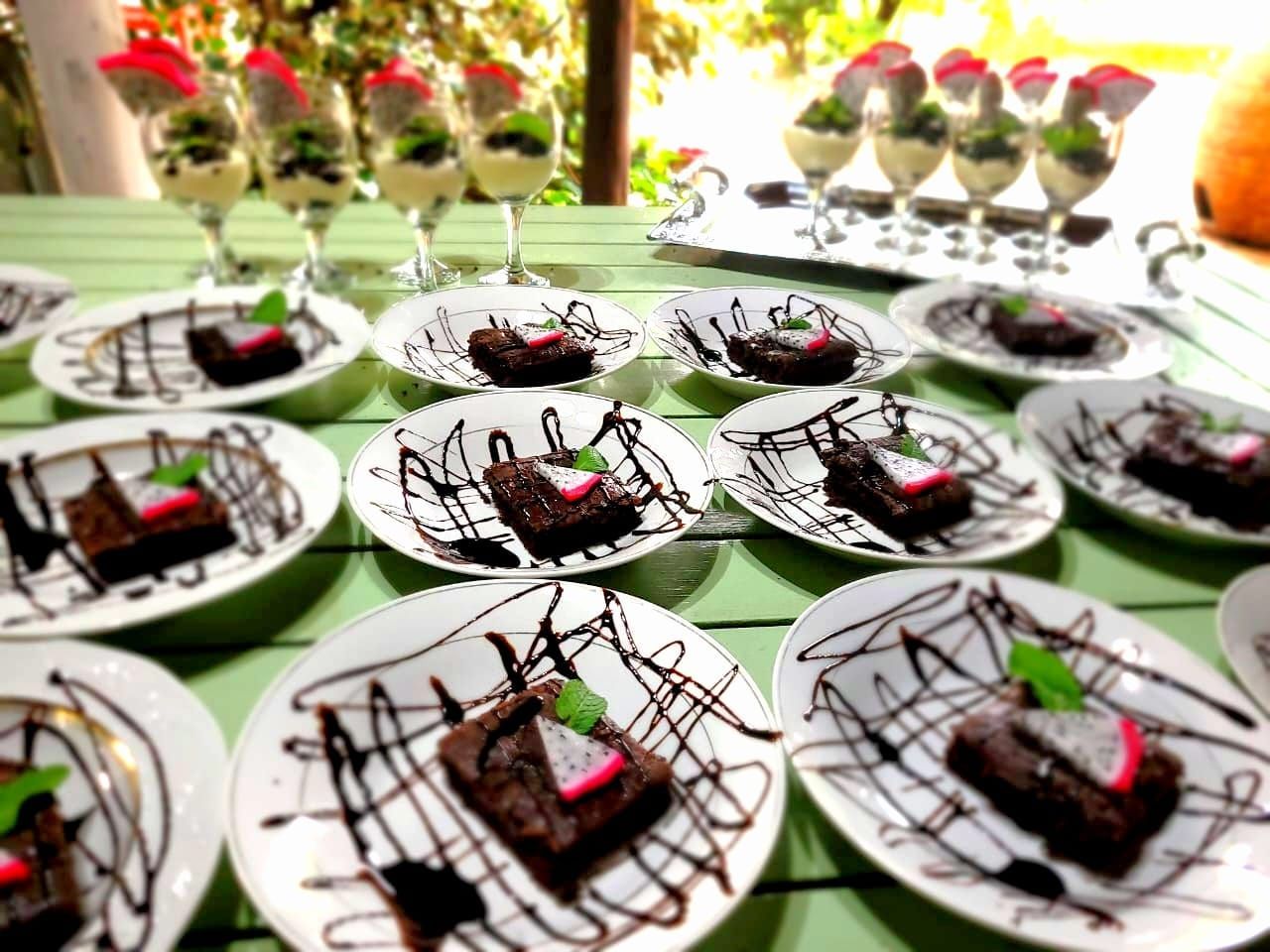 Dessert plates with chocolate-drizzled brownies and strawberries on a green tablecloth, with flower vases in back