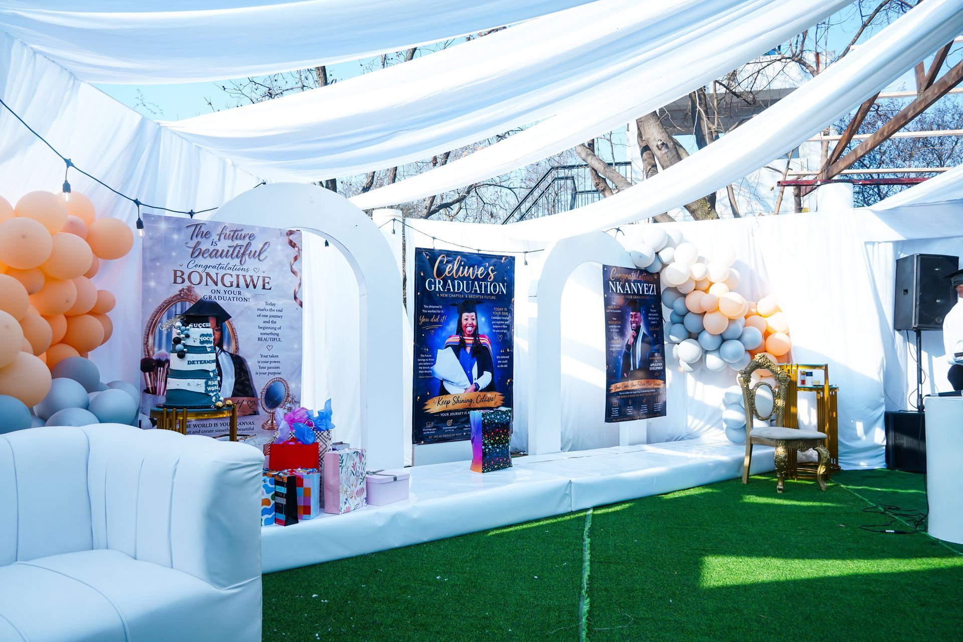 White outdoor event tent with balloons, posters, and a white couch on green turf under draped fabric.