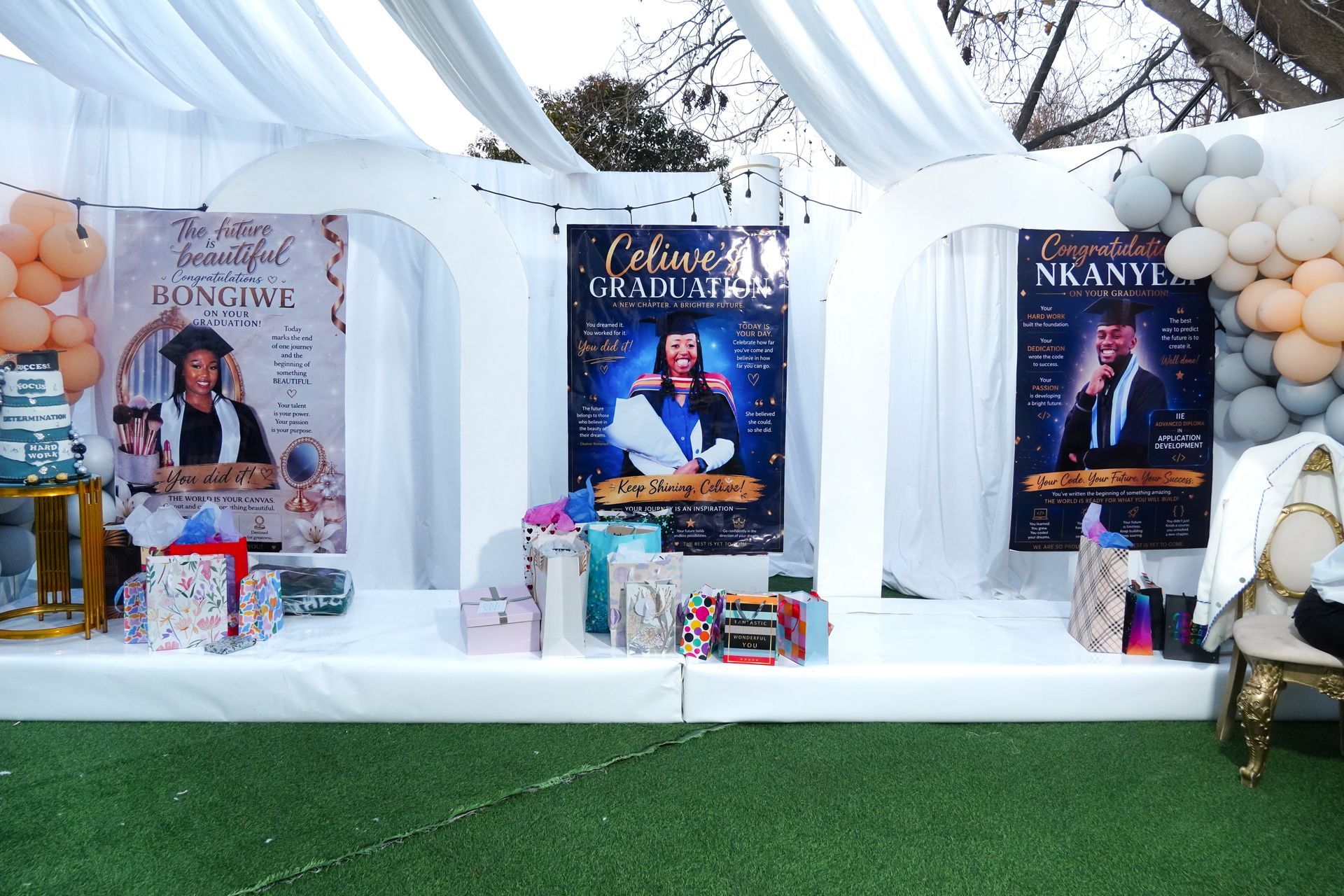 Outdoor event booth with three large posters, wrapped gifts, white drapes, balloons, and green turf backdrop