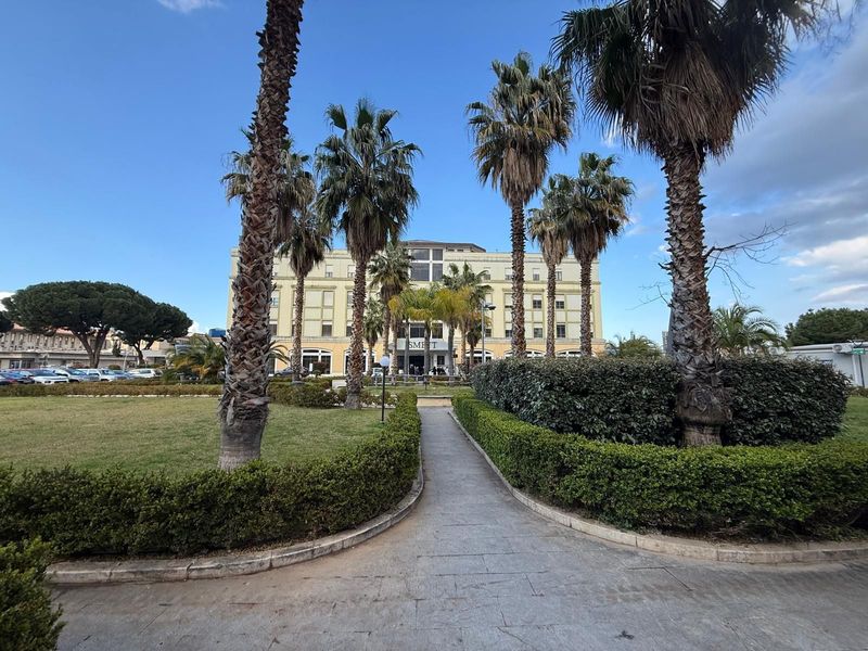 Un sentiero lastricato attraversa un parco e conduce a un grande edificio beige, fiancheggiato da siepi ben curate e alte palme.
