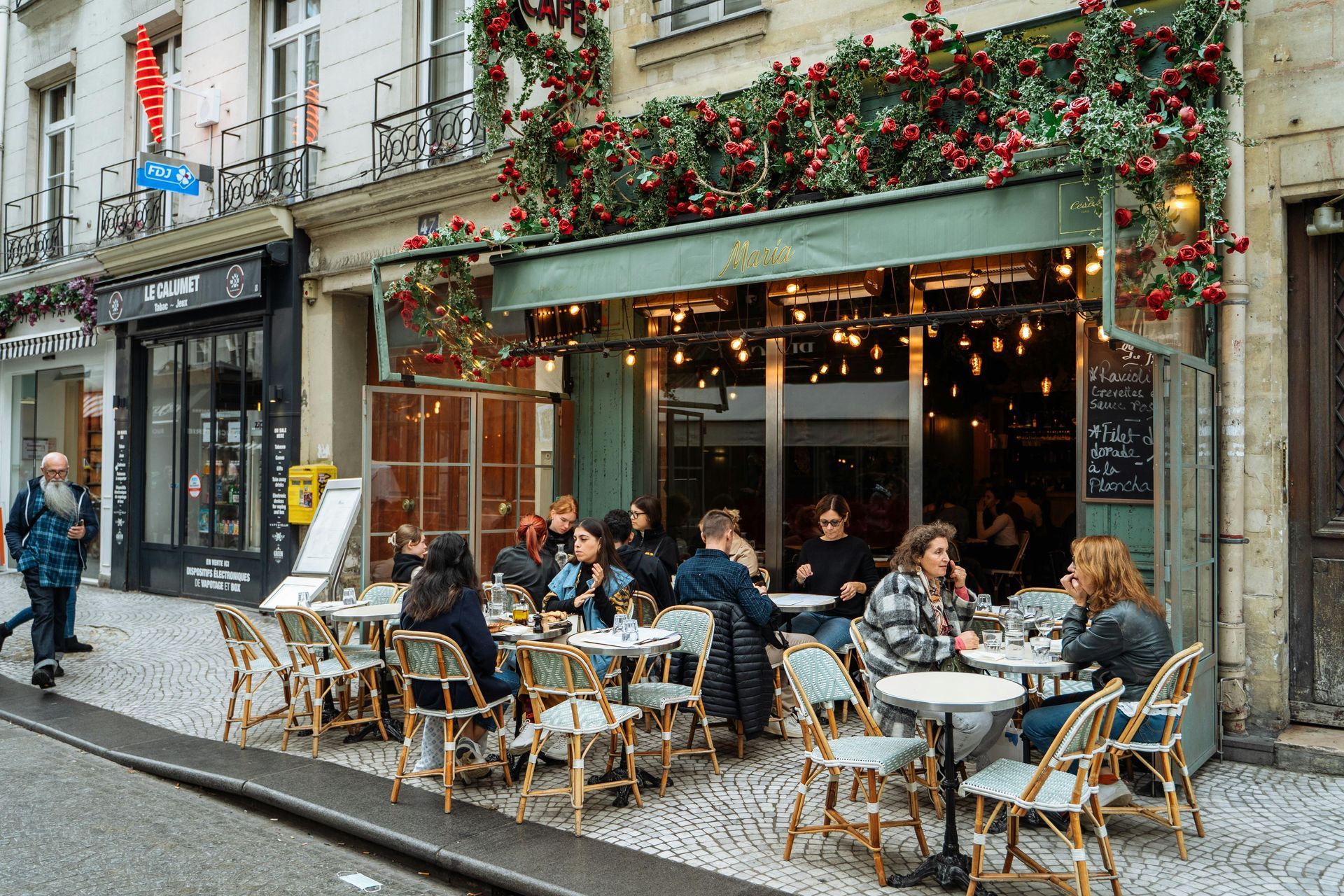Outdoor café with diners seated at wicker tables along a cobblestone street, under flower-covered awnings.