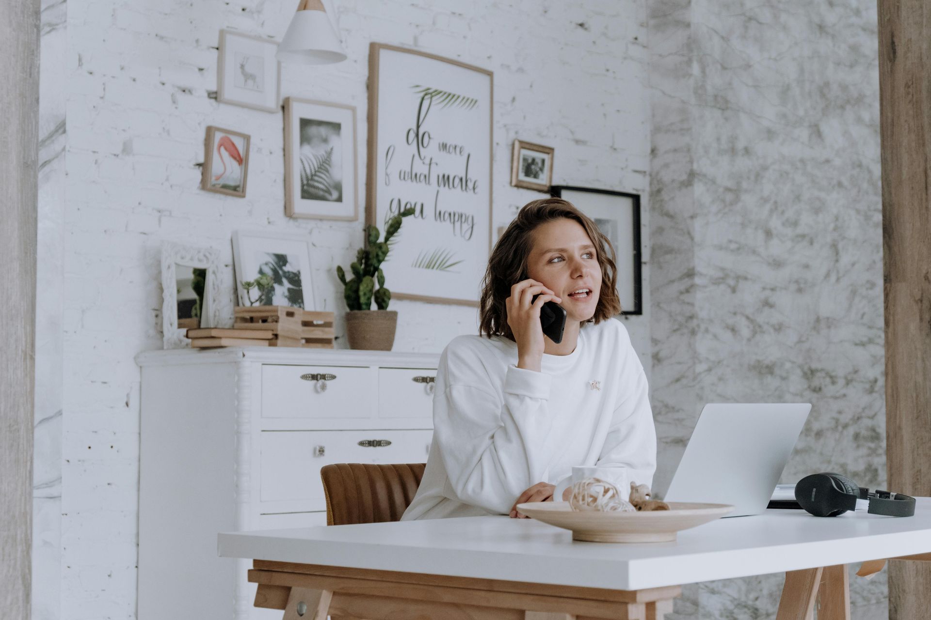 Woman on the phone in a home office with a laptop and artwork on the wall.