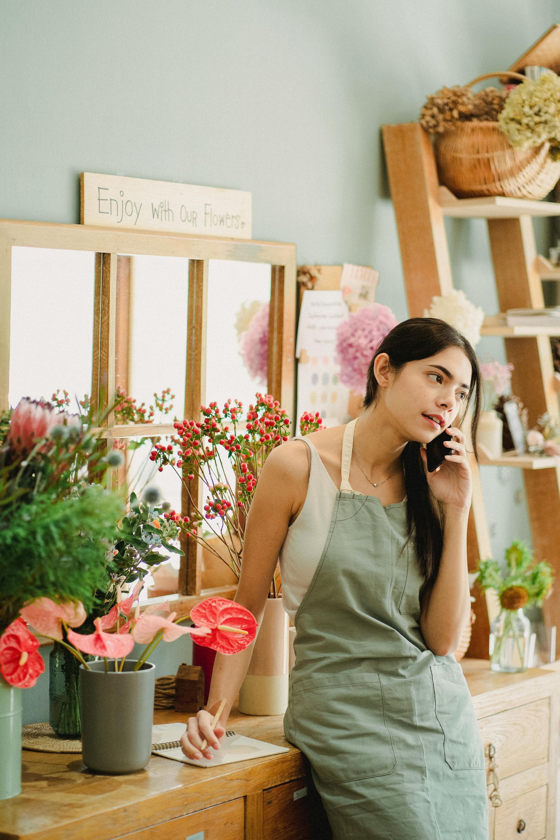Woman in green apron holding a phone in a bright flower shop, surrounded by colorful bouquets.