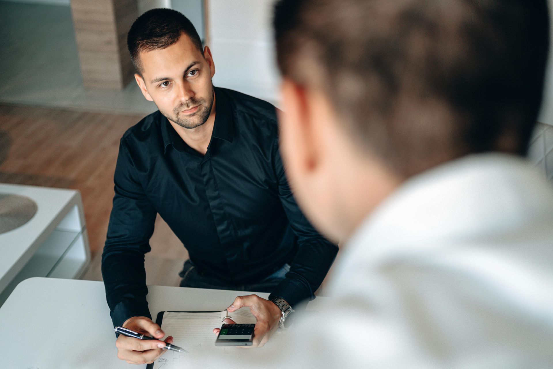 Man in dark shirt taking notes during a meeting with another person in a white shirt