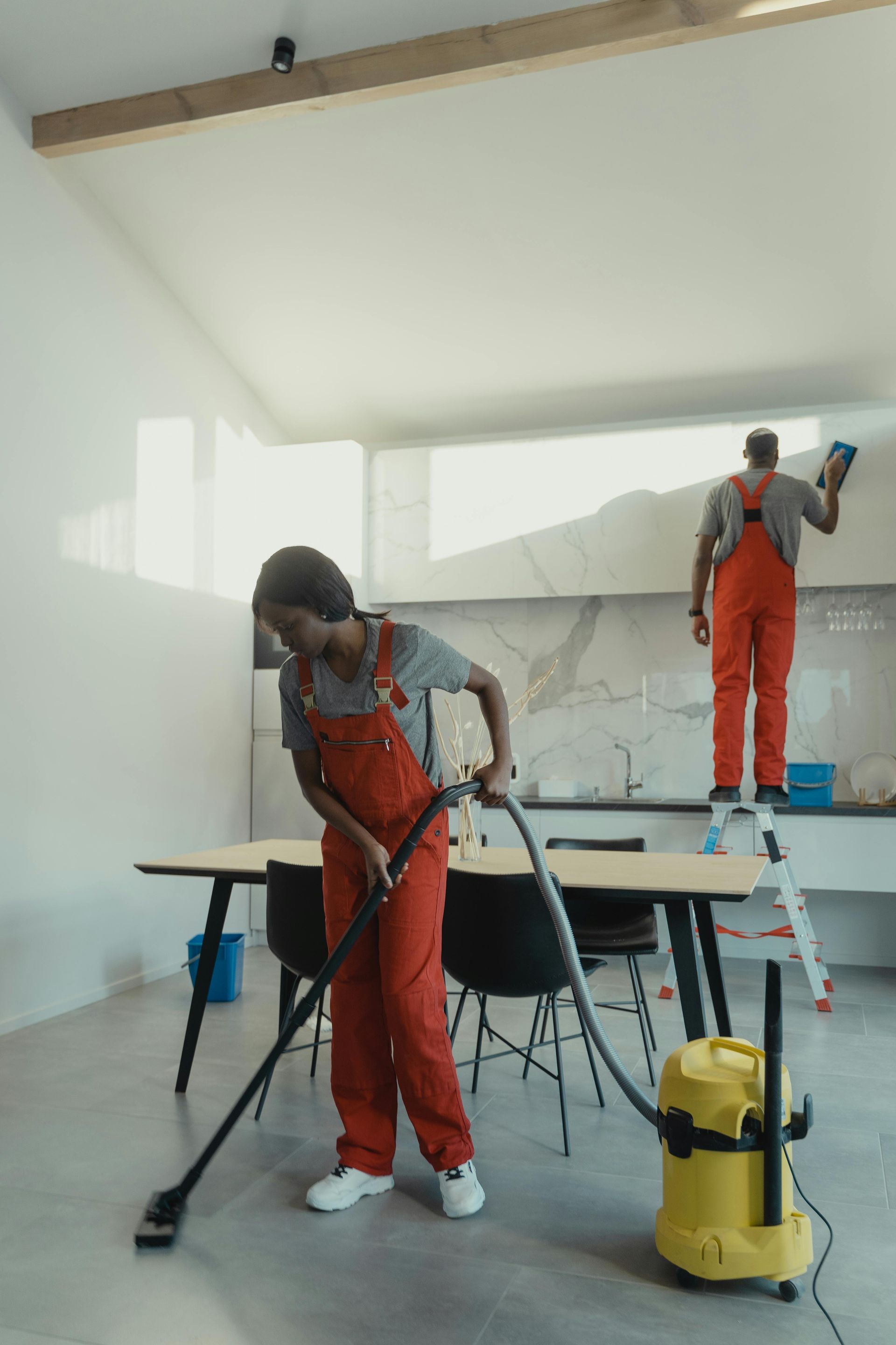 Two workers in orange overalls painting a bright white room with rollers and a yellow vacuum nearby