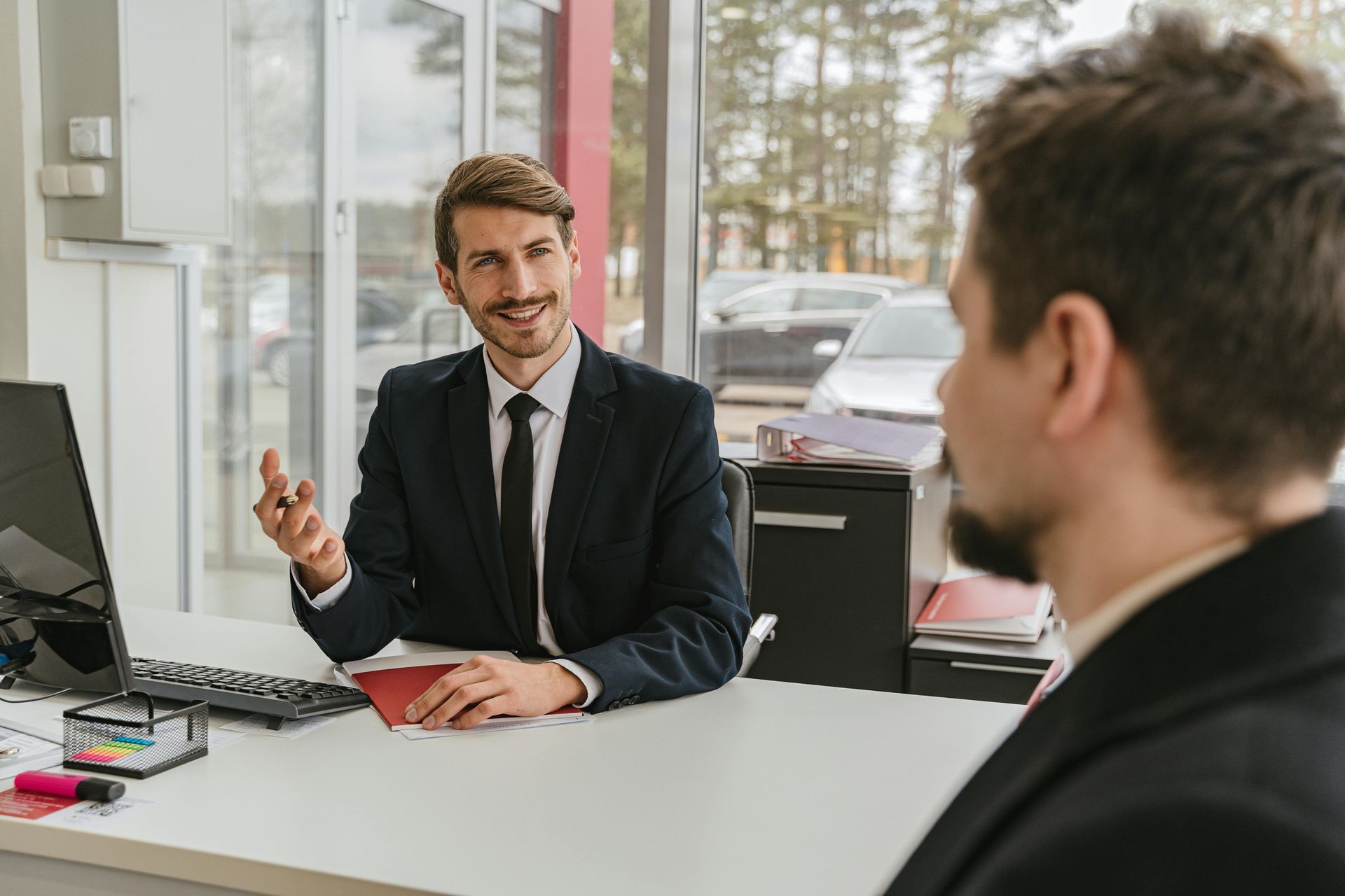 Two men talking in an office, one seated at a desk with a computer and gesturing while the other listens.