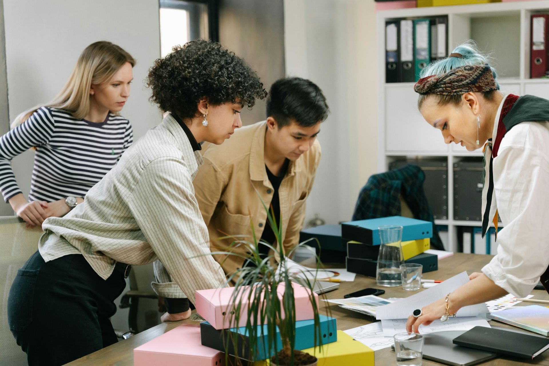 Four coworkers lean over a desk, reviewing papers and a laptop in a bright office.
