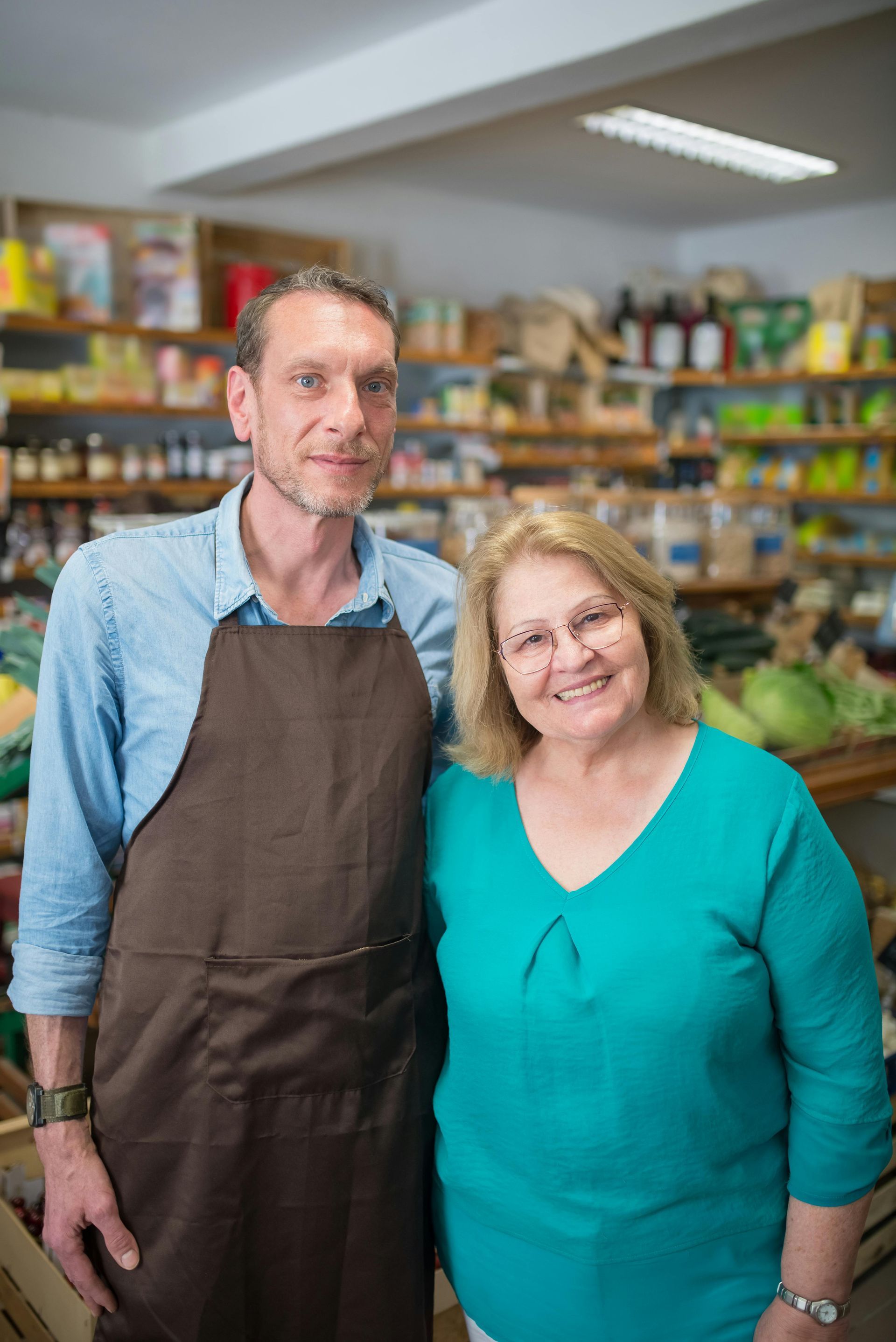Two shop staff in aprons standing inside a colorful grocery store, smiling at the camera.