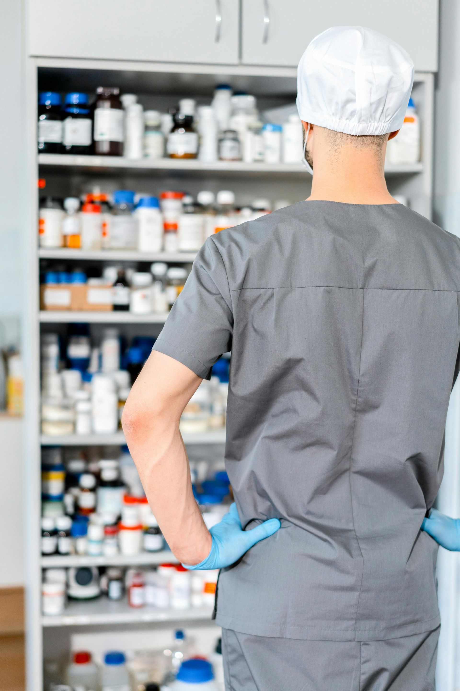 Healthcare worker in gray scrubs and blue gloves, seen from behind, facing medicine shelves in a pharmacy.