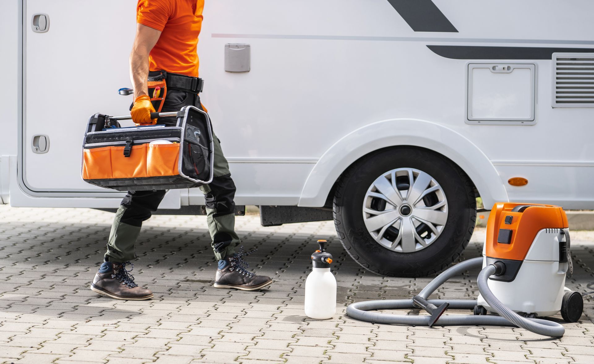 Person with toolbox, walks by RV, near vacuum and cleaning bottle.