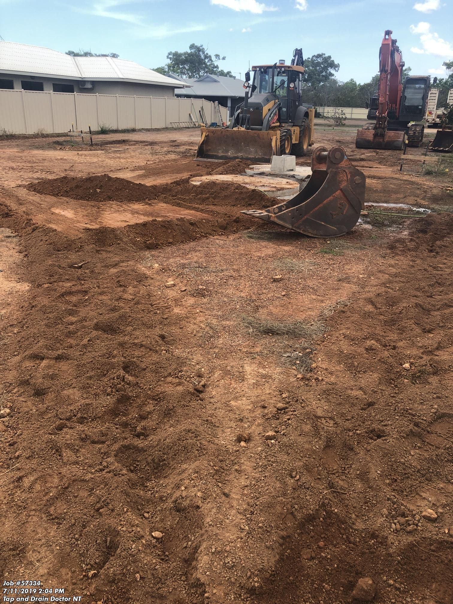 Construction site with an excavator digging a dirt trench beside another machine and a long white building.