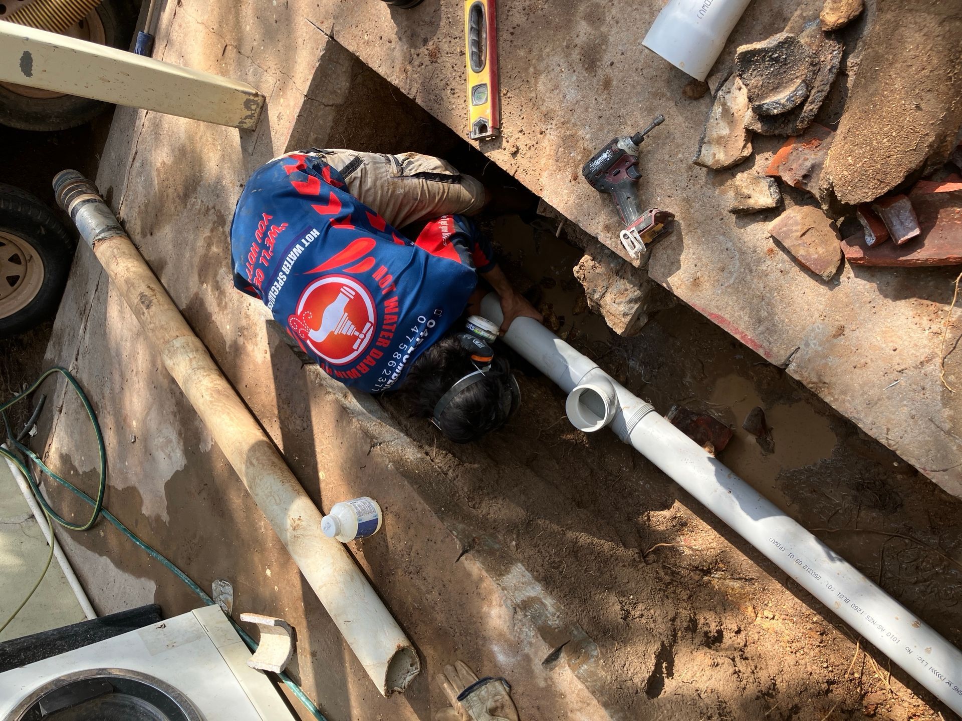 Worker in blue and red shirt repairing white PVC pipes in a dirt trench with bricks and debris nearby — Tap & Drain Doctor NT in Winnellie, NT