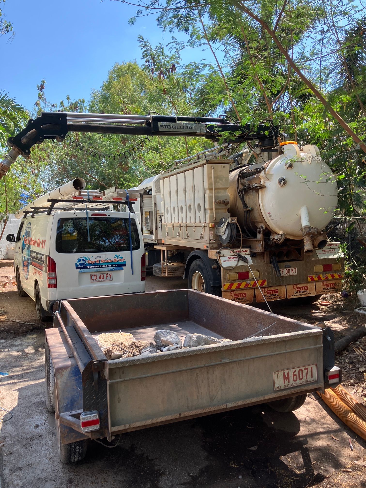 Utility truck and vacuum tanker on a dirt road, with a trailer in the foreground and trees overhead — Tap & Drain Doctor NT in Winnellie, NT