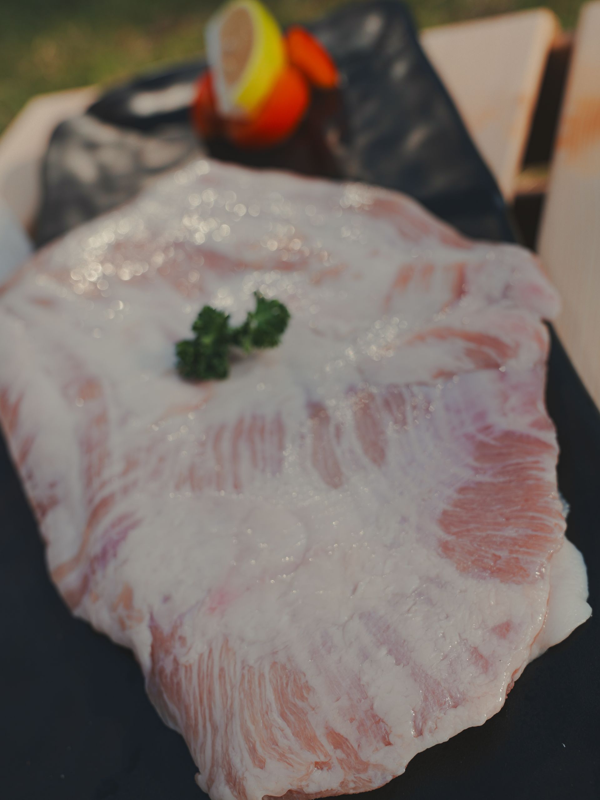 一塊生的大理石紋豬肉，配上一小枝歐芹，放在黑色盤子上，背景是檸檬和番茄。