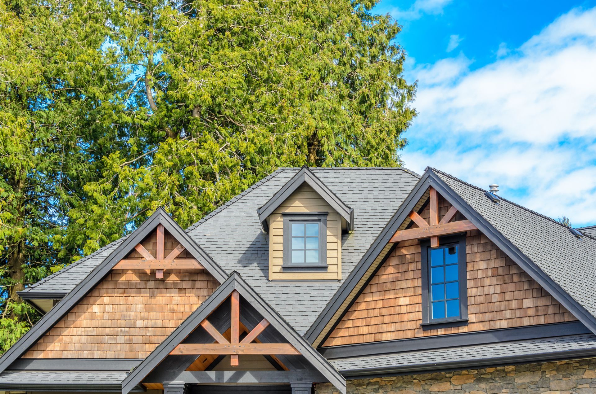 House roof with gables, dormers, and wood accents, against a green tree and blue sky.