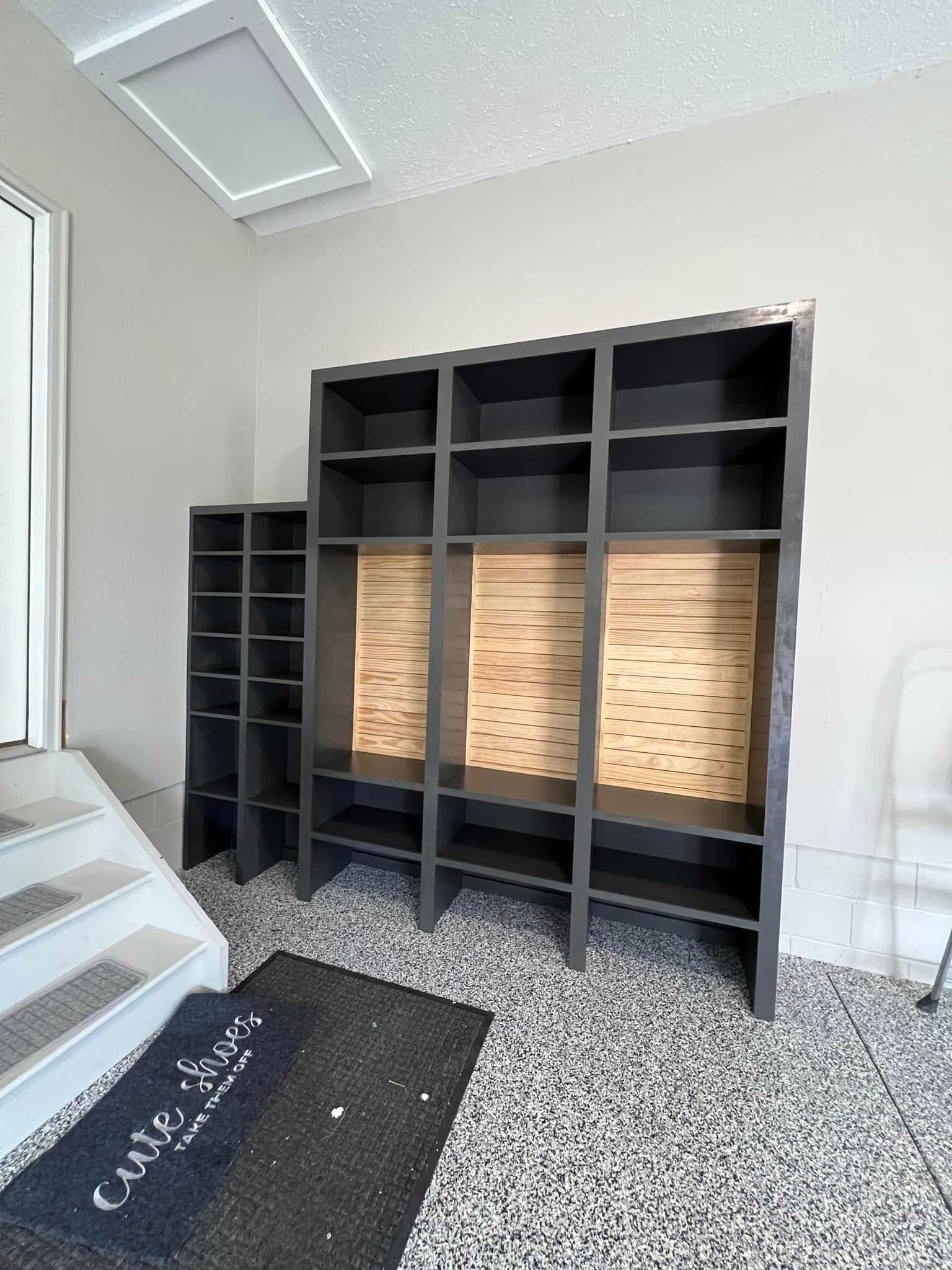 Dark gray built-in shelving unit with wood backing, next to a narrow shoe rack. Mat on speckled floor.
