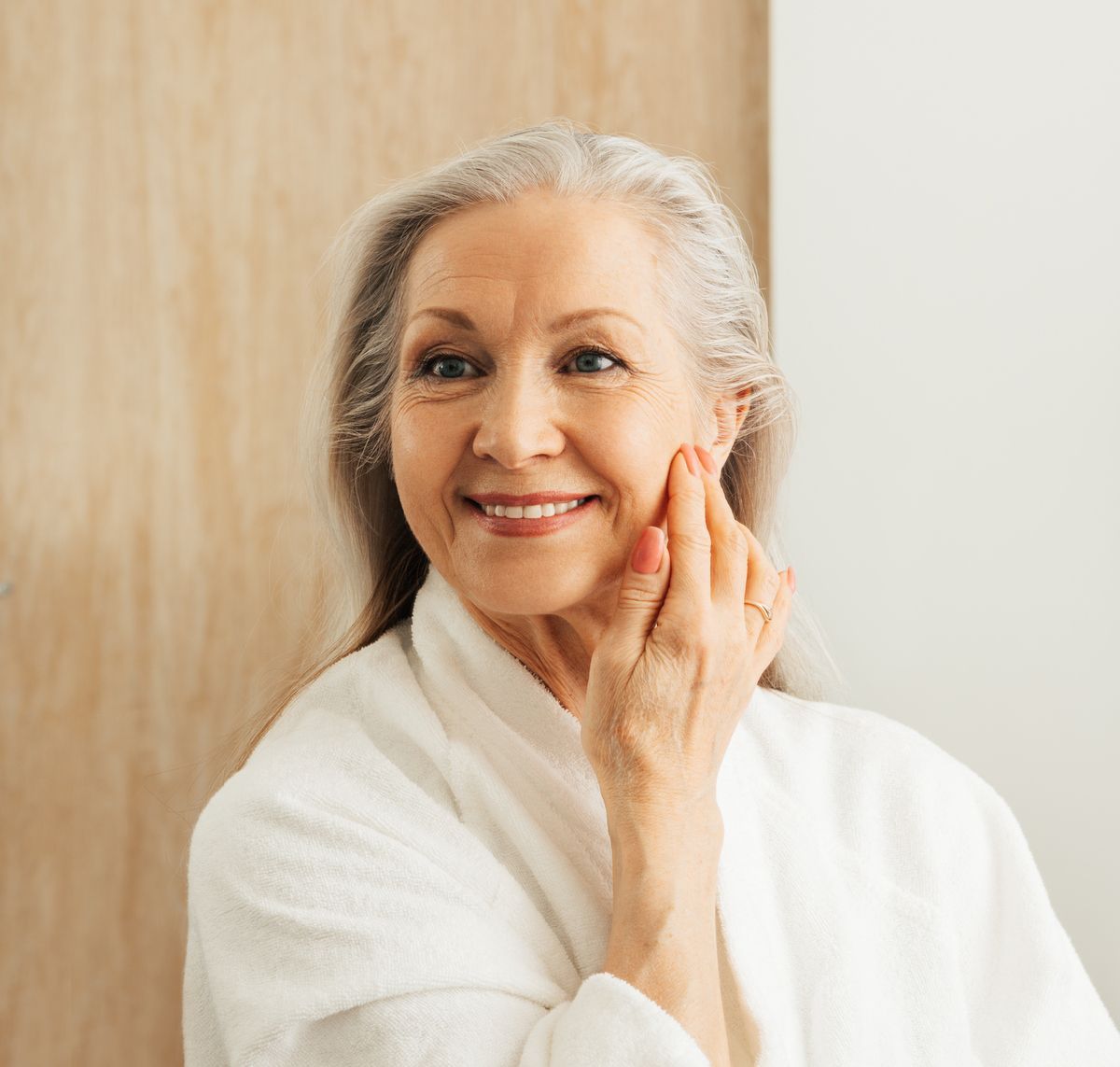 Smiling woman in a white robe touching her face in a bright bathroom