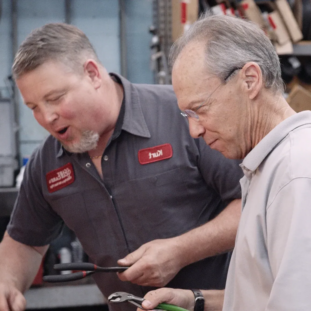 Two men examining tools in a shop. One points, the other looks on. Gray shirts, indoor setting.
