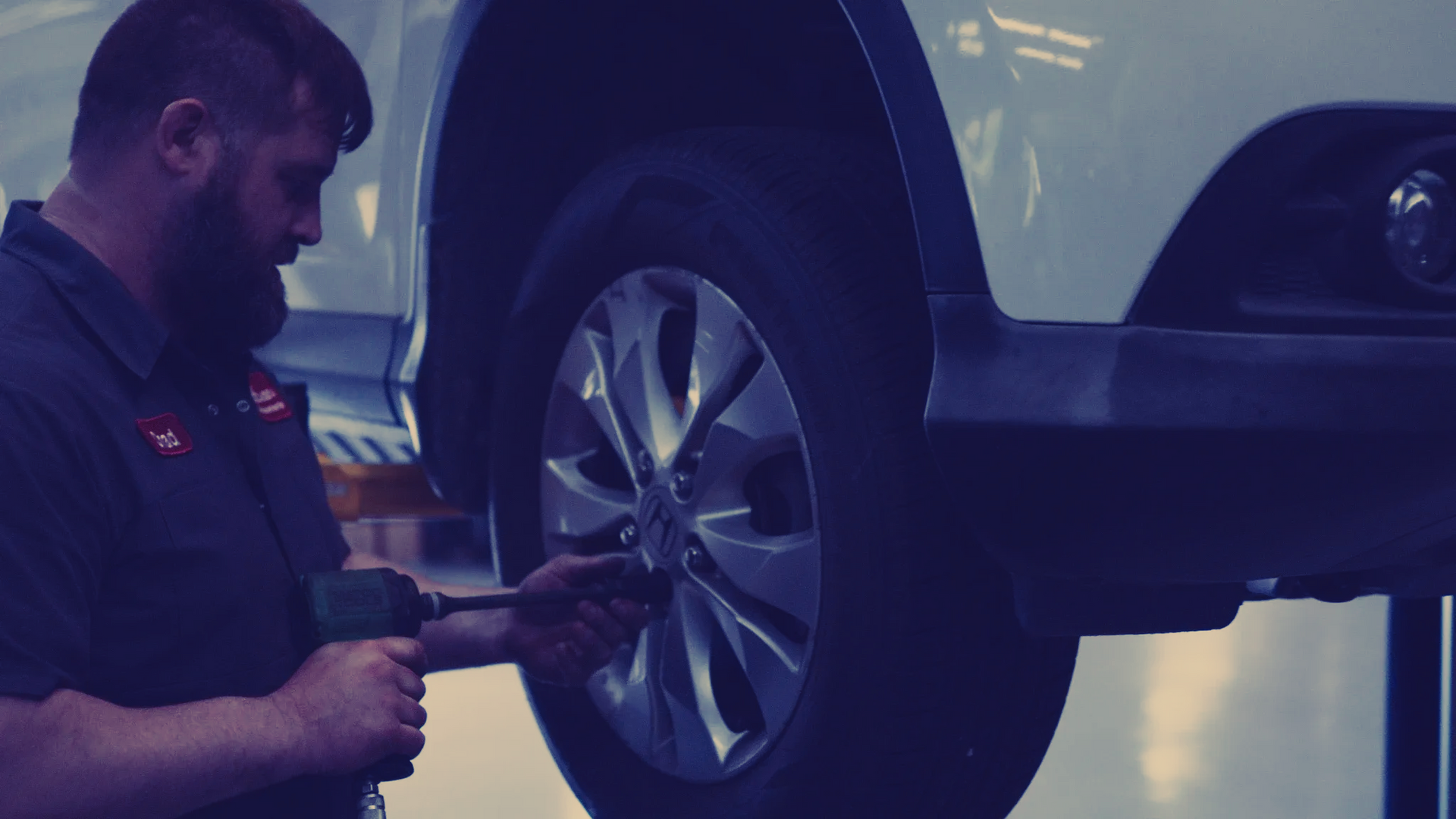 Mechanic using a power tool to remove a car tire. Garage setting; vehicle on a lift.