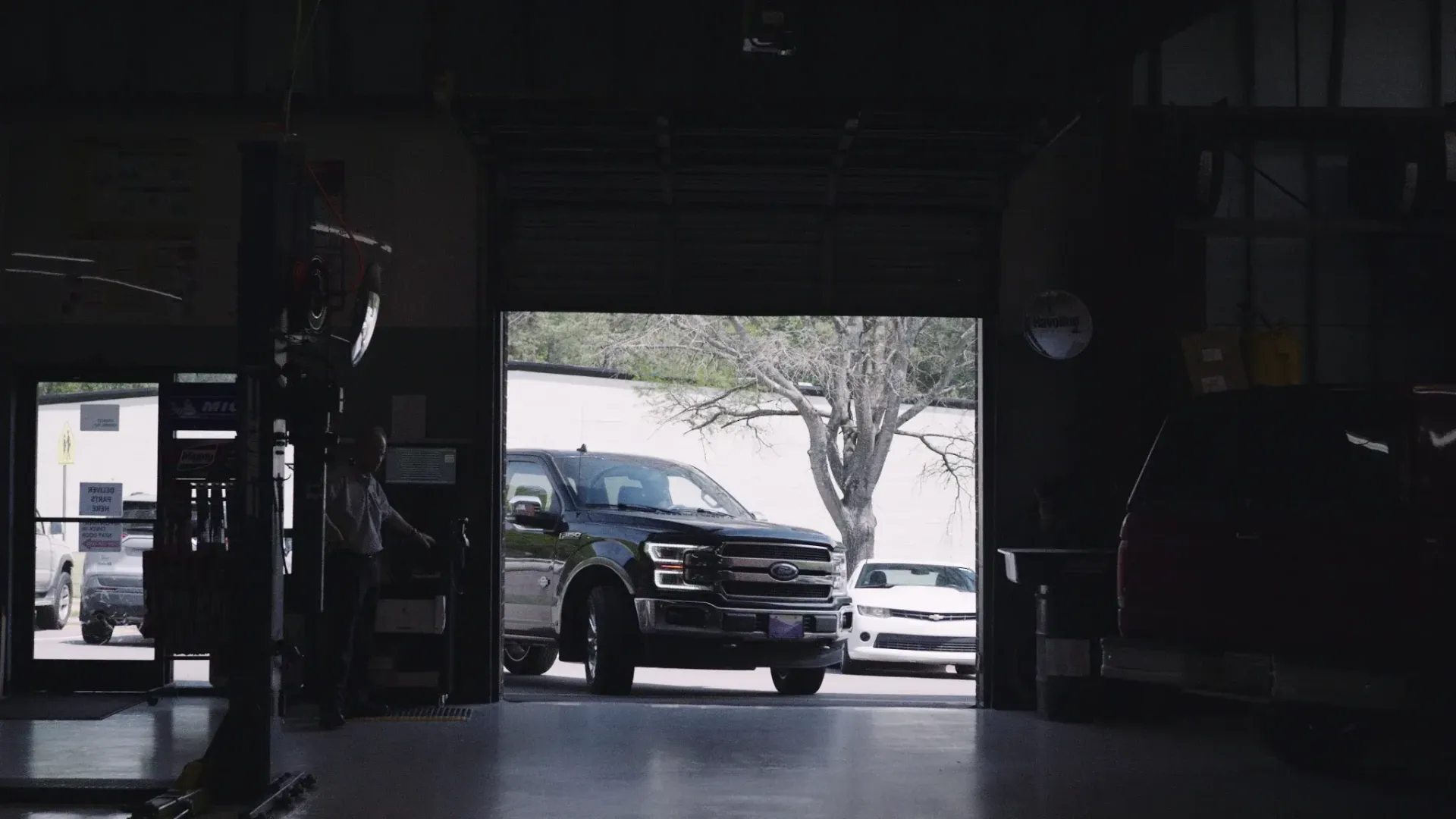 Dark garage with a black pickup truck and white sports car near the open door, trees visible outside.