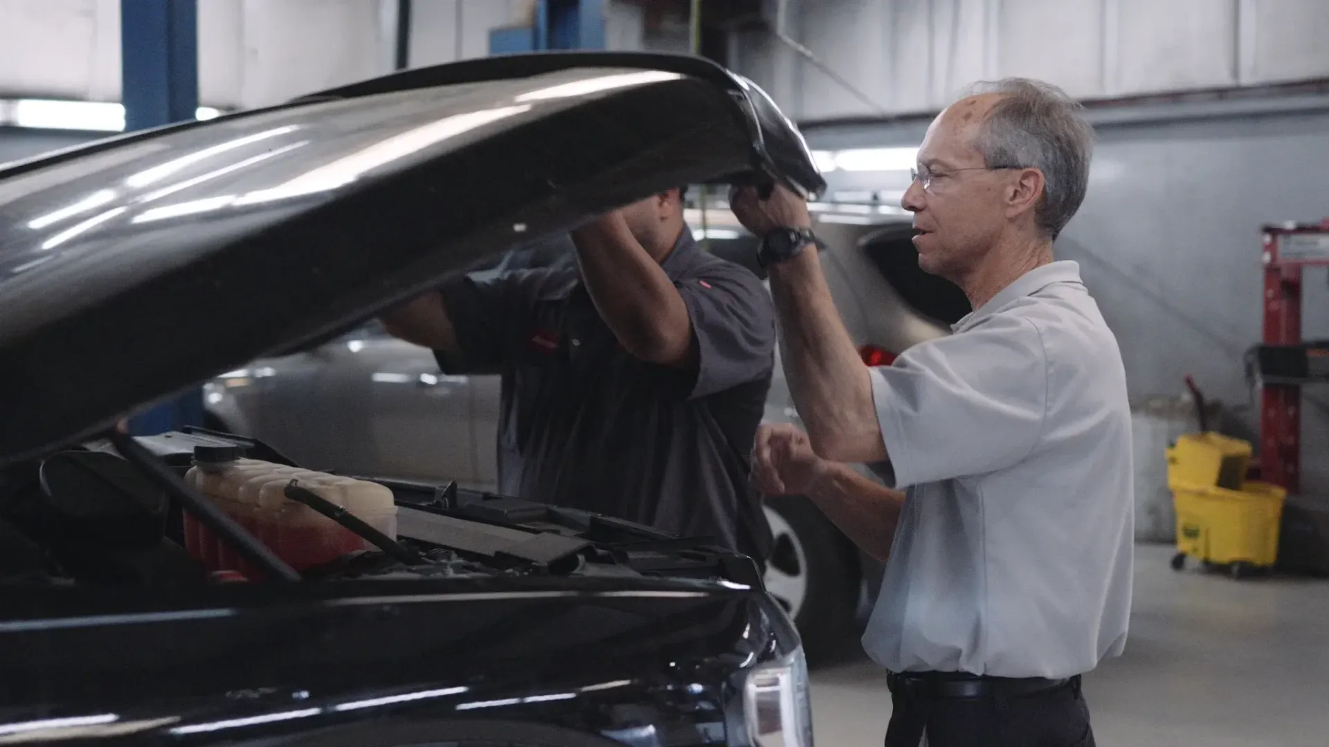 Two people opening a car hood in a repair shop.