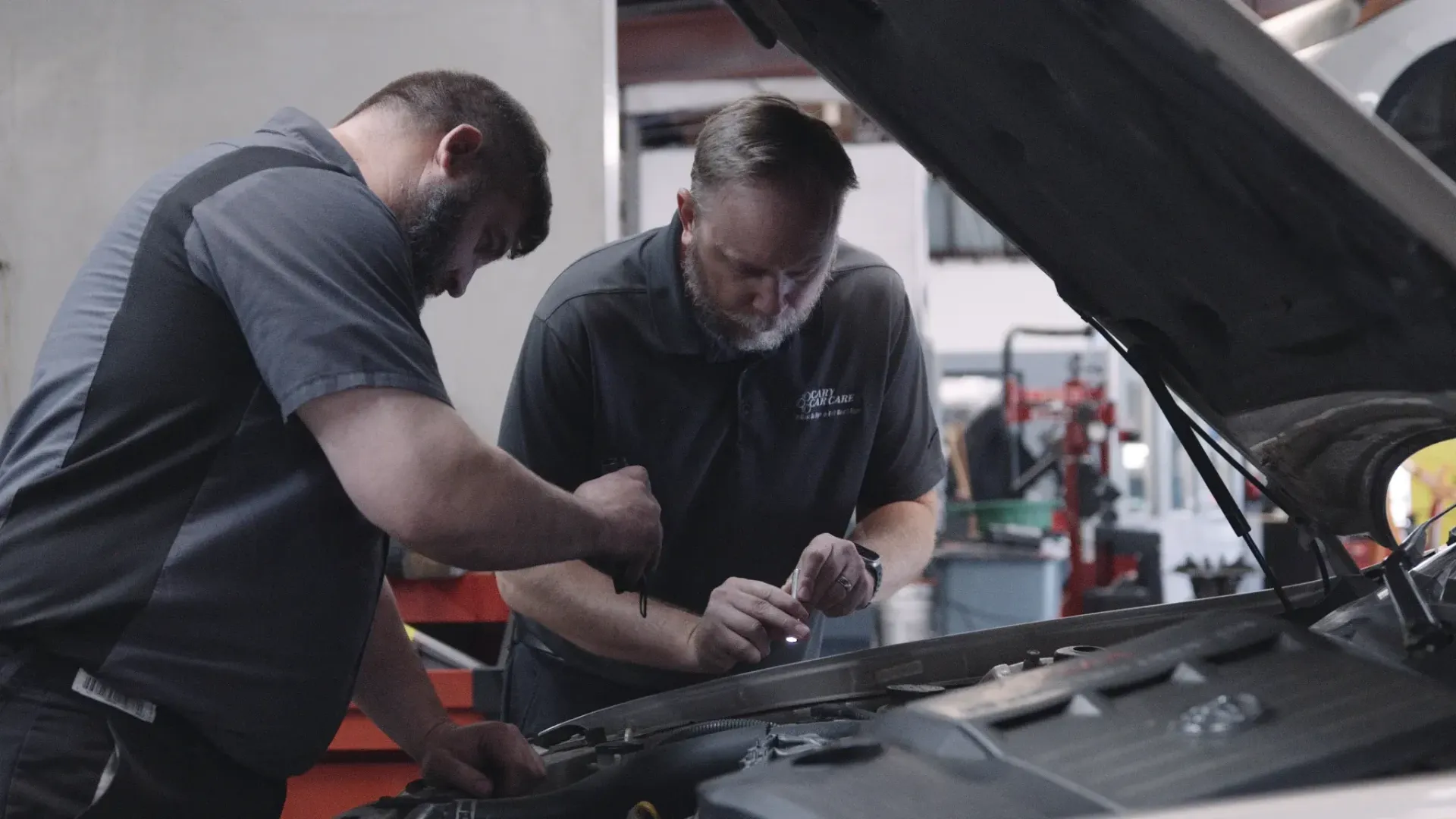 Two mechanics in a garage working on a car engine with tools; gray and black uniforms, looking down.