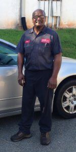 A man in a mechanic's uniform stands in front of a car. He's wearing glasses and appears to be smiling.