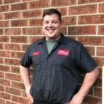 Man in work uniform smiling, leaning against a brick wall.