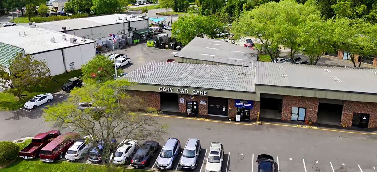 Aerial view of a commercial building with a parking lot and several vehicles. Green trees surround the buildings.