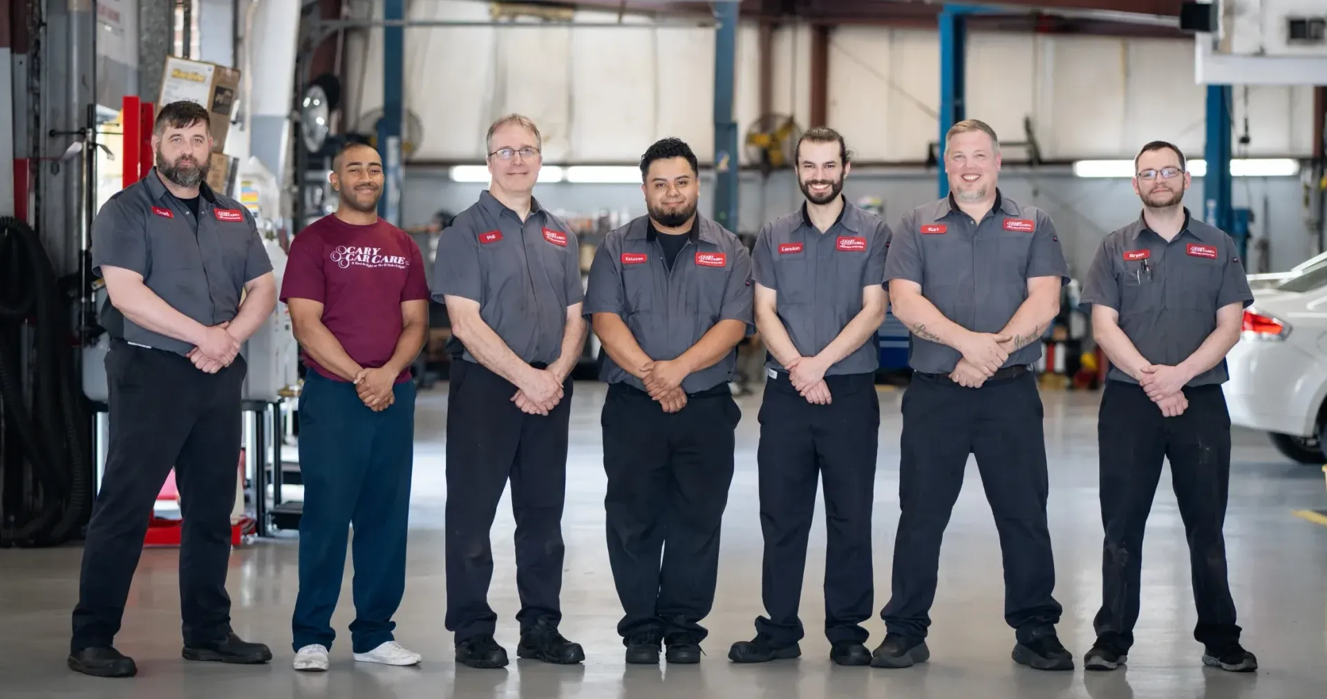 Seven auto mechanics in gray work shirts stand shoulder to shoulder in a garage, arms crossed.