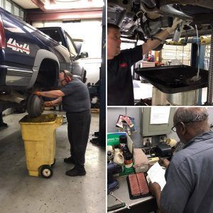 Mechanics working on cars in a garage. One changes a tire, another drains oil, and the last reviews paperwork.