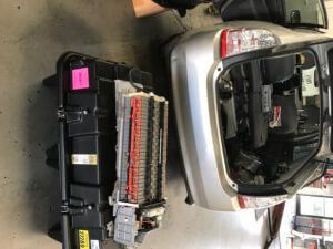 Hybrid car with open trunk and removed battery pack. Silver car, black battery, repair shop setting.