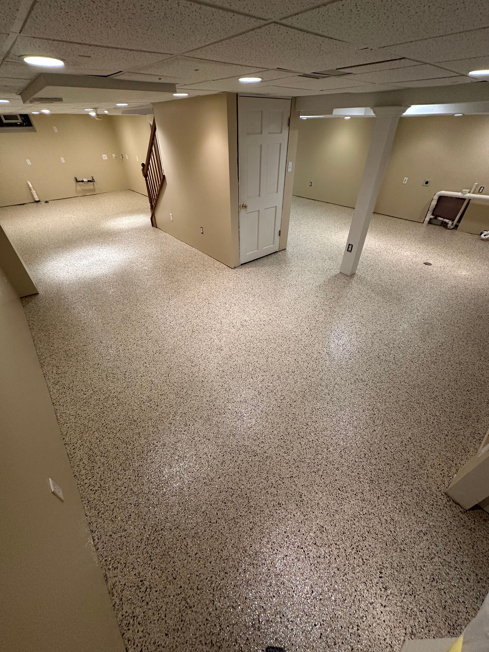 Empty basement room with speckled floor, beige walls, support columns, and overhead fluorescent lights