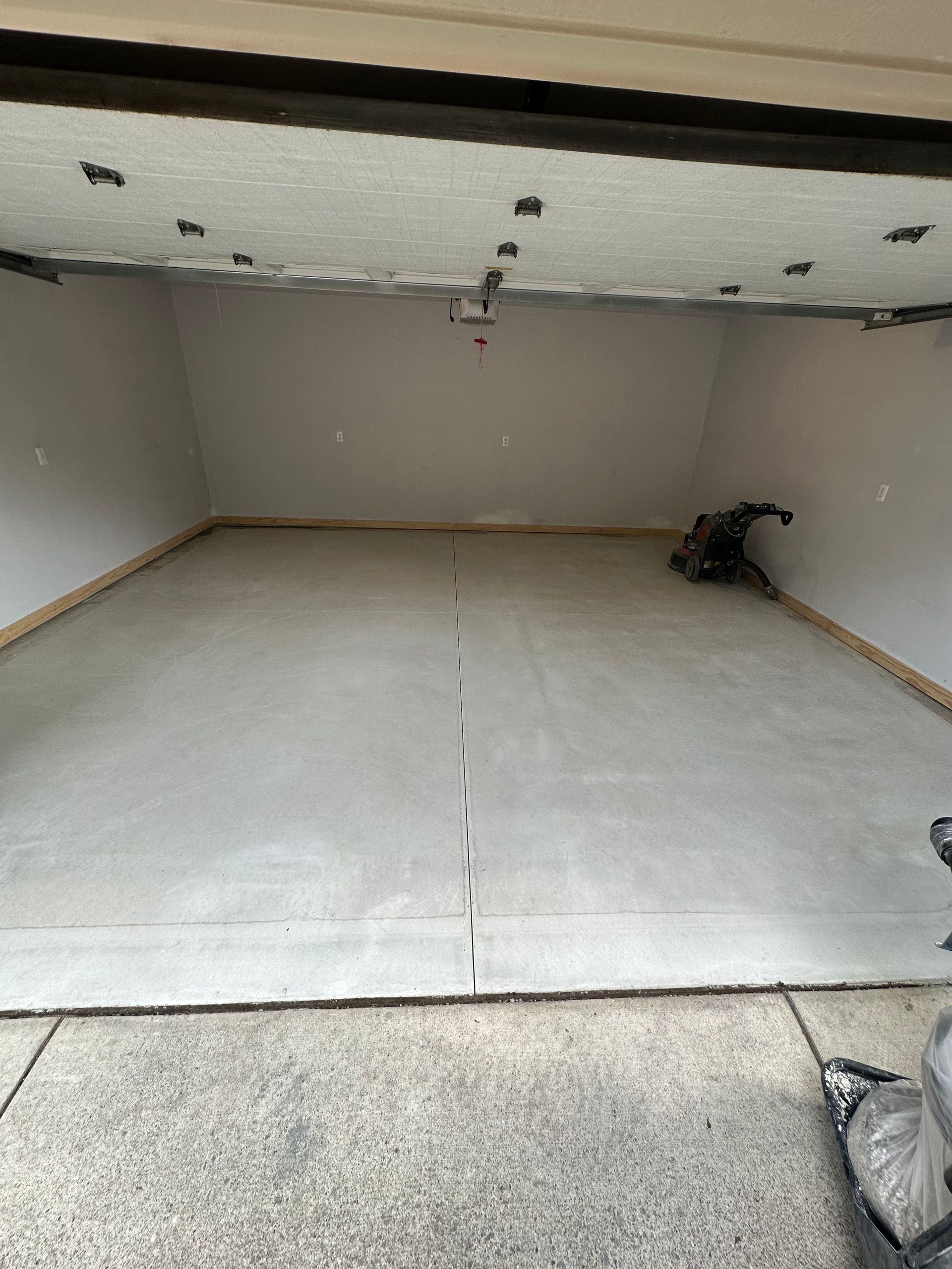 Empty garage with white walls and floor, exposed ceiling, and a tarp or bag in the right corner.