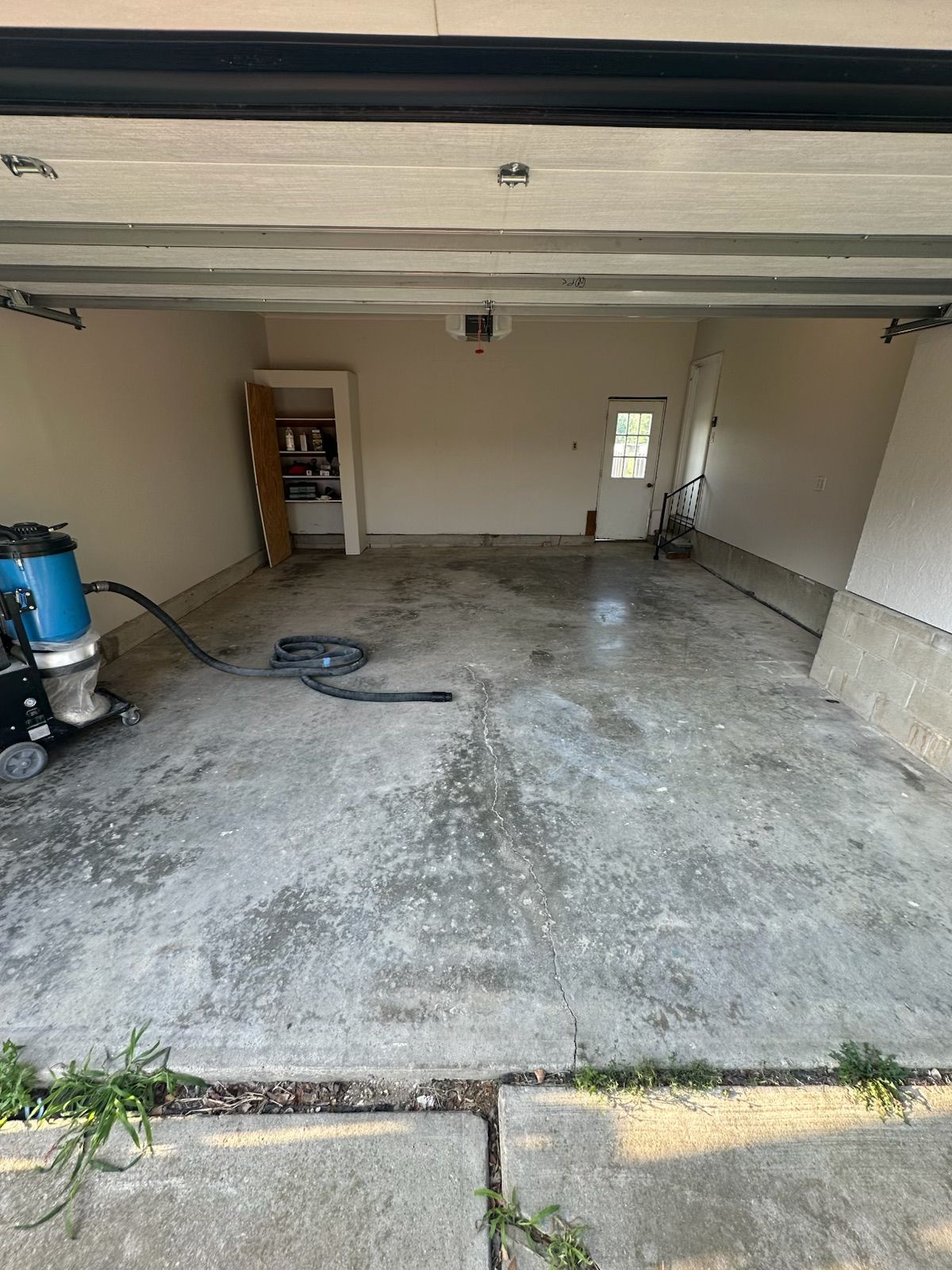 Empty garage with bare concrete floor, open door, shelves, and a blue barrel on the left.