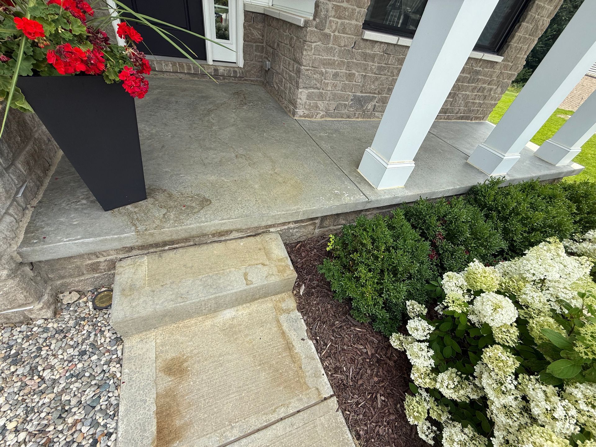 Front porch with short concrete steps, white columns, and flower planters beside a gravel walkway.