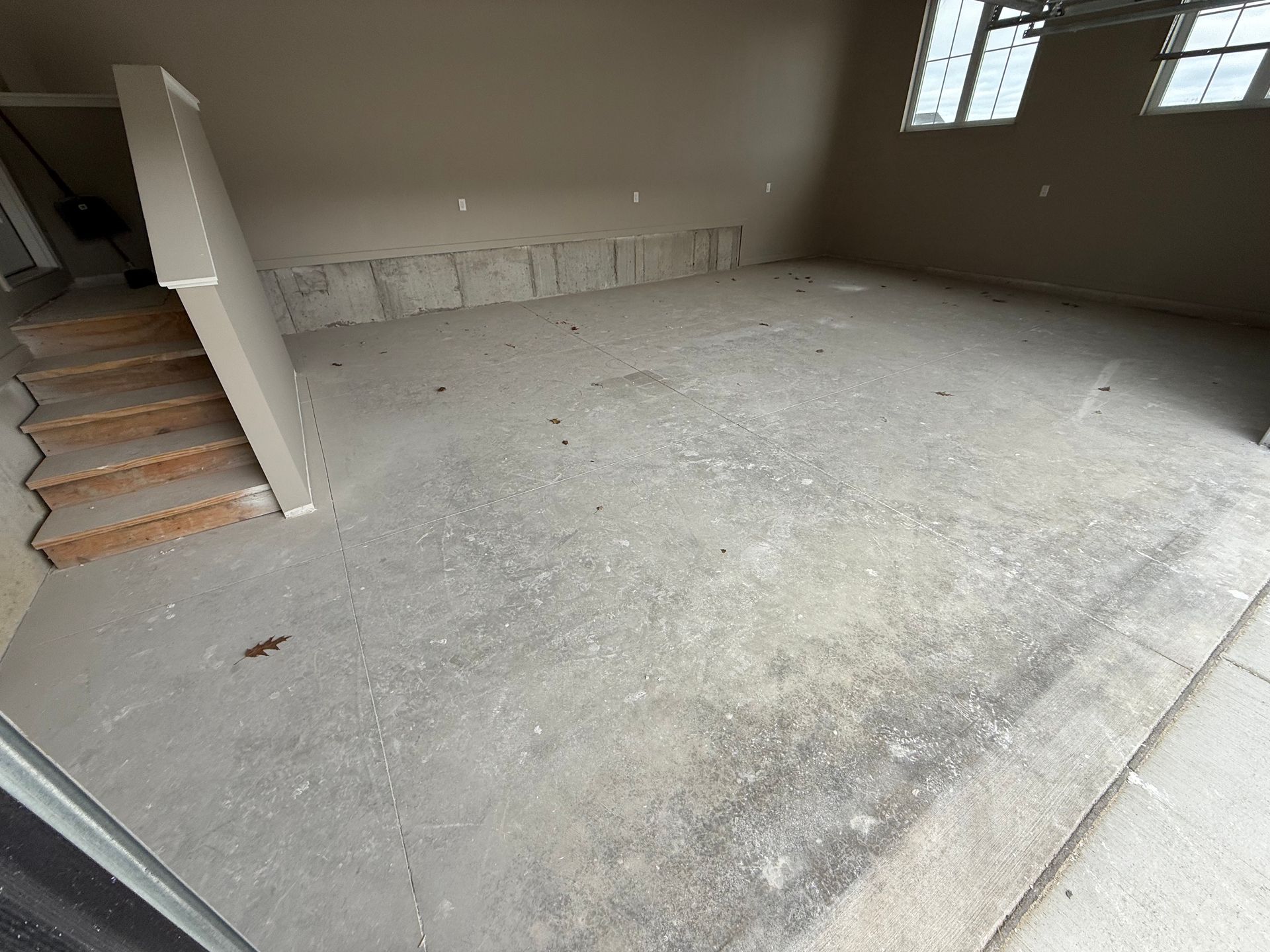 Empty unfinished room with bare concrete floor, beige walls, a staircase on the left, and a window on the right.