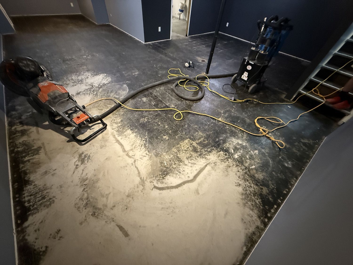 Wet floor being resurfaced with a floor grinder, dust spread across a room with cords and tools.
