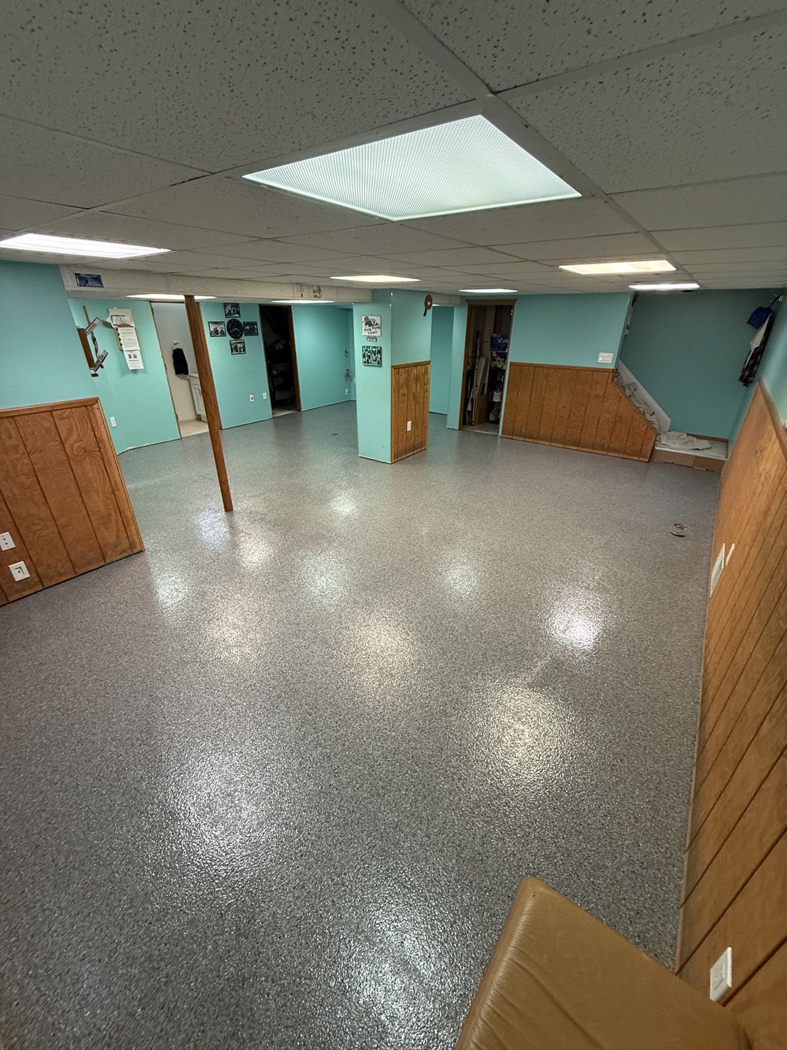 Empty teal room with speckled floor, wood-paneled walls, and fluorescent ceiling lights