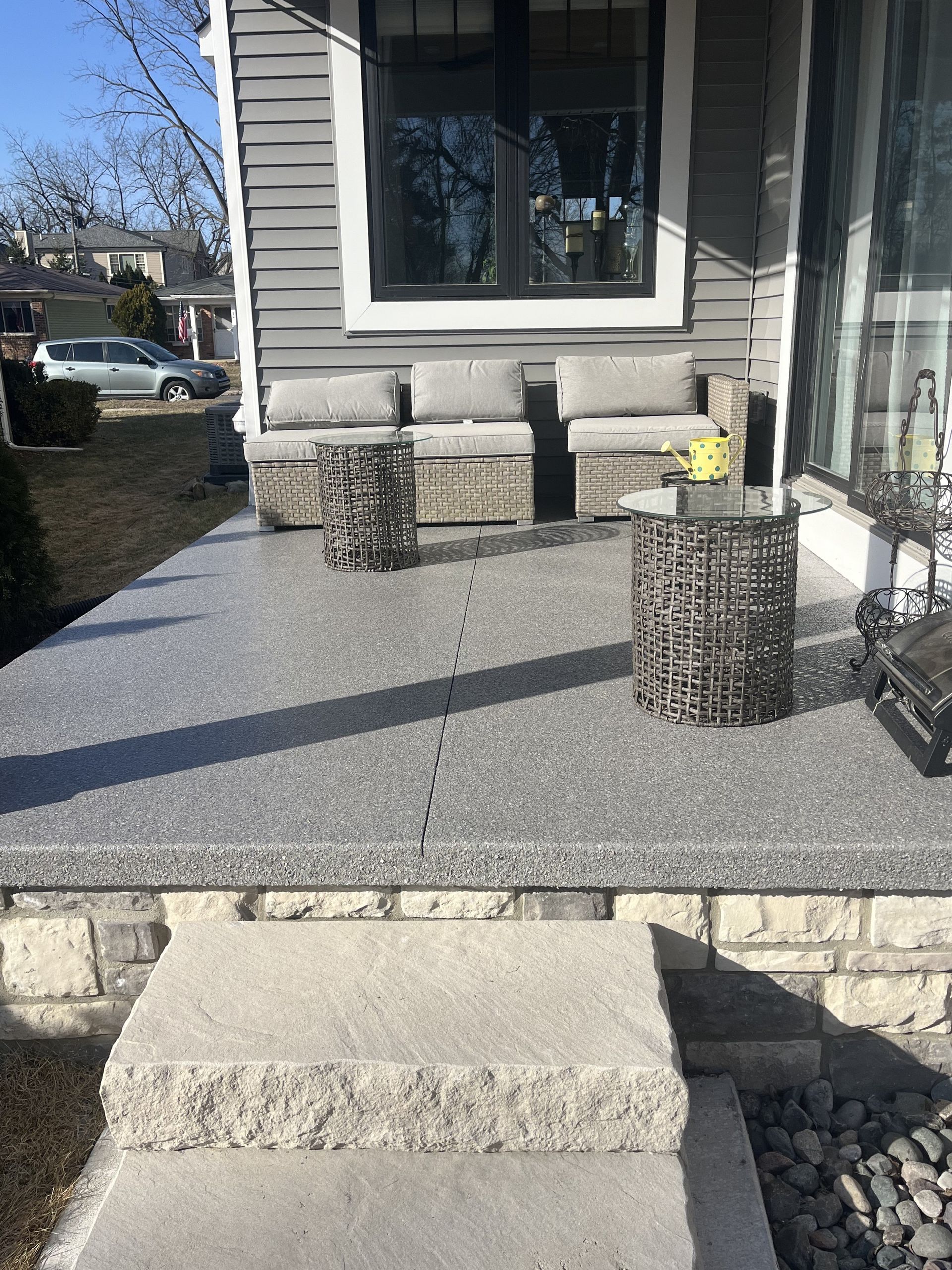 Front porch with concrete steps, pebble-finished patio, and wicker seating outside a gray house
