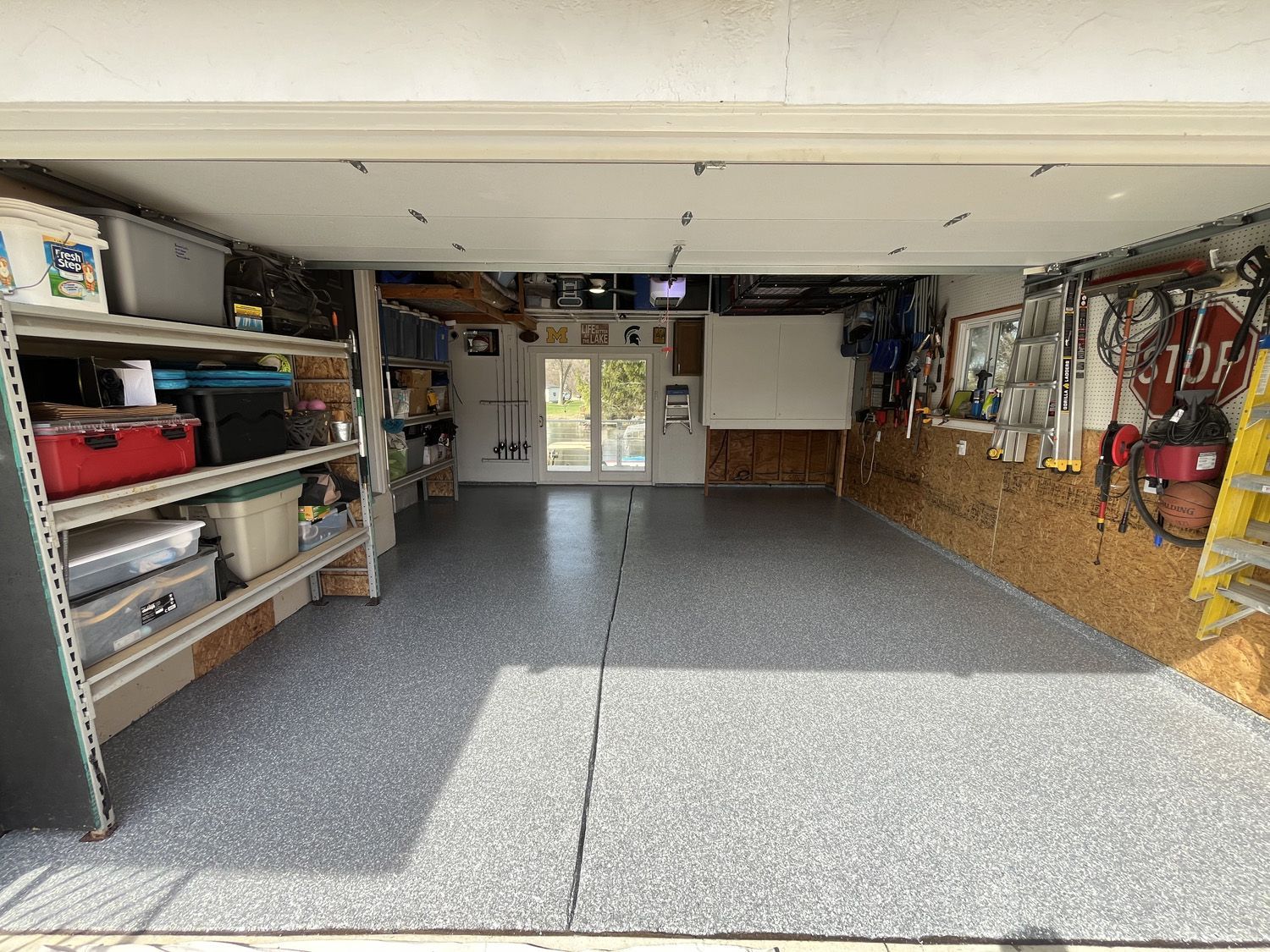 Empty garage interior with gray speckled floor, shelves on the left, and tools and storage along the walls.