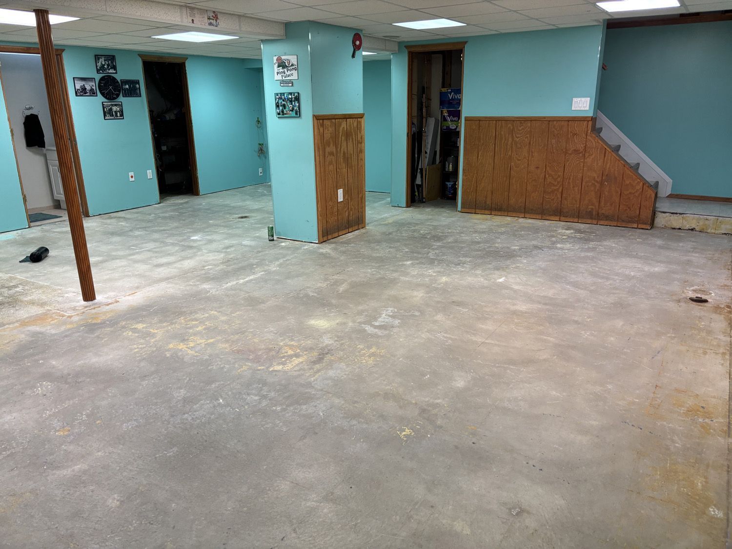 Empty basement room with bare concrete floor, teal walls, wood paneling, and stairs on the right.