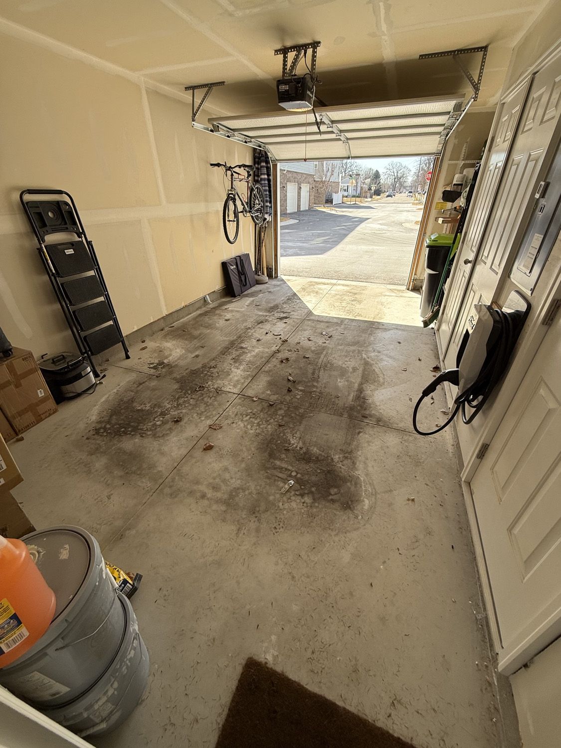 Garage interior with open door, a ladder, tools, and a view outside to a driveway and trees.