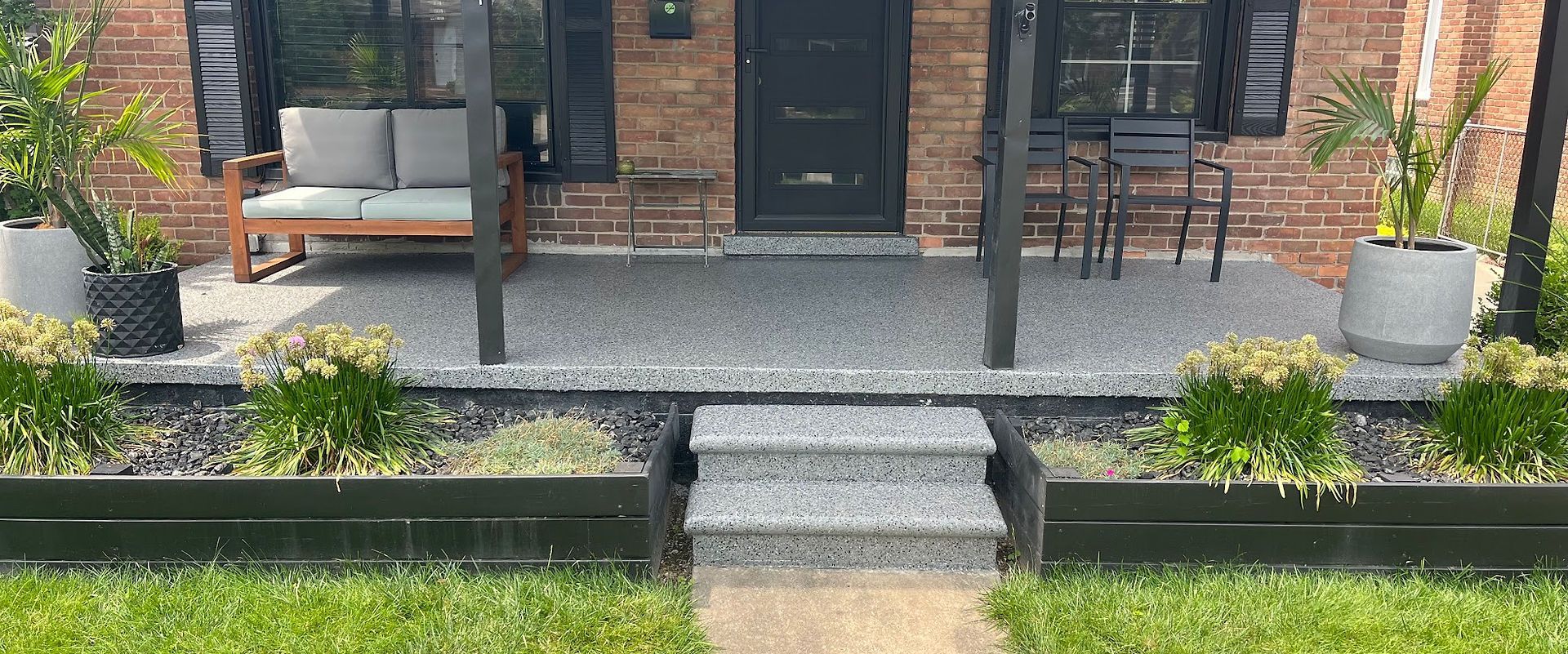 Front porch with gray steps, black door, brick house, bench seating, and potted plants.