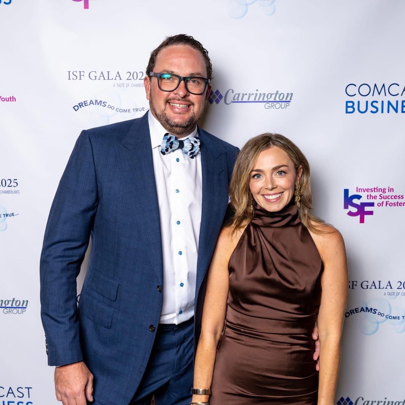 Two people posing on a gala red carpet before a branded step-and-repeat backdrop